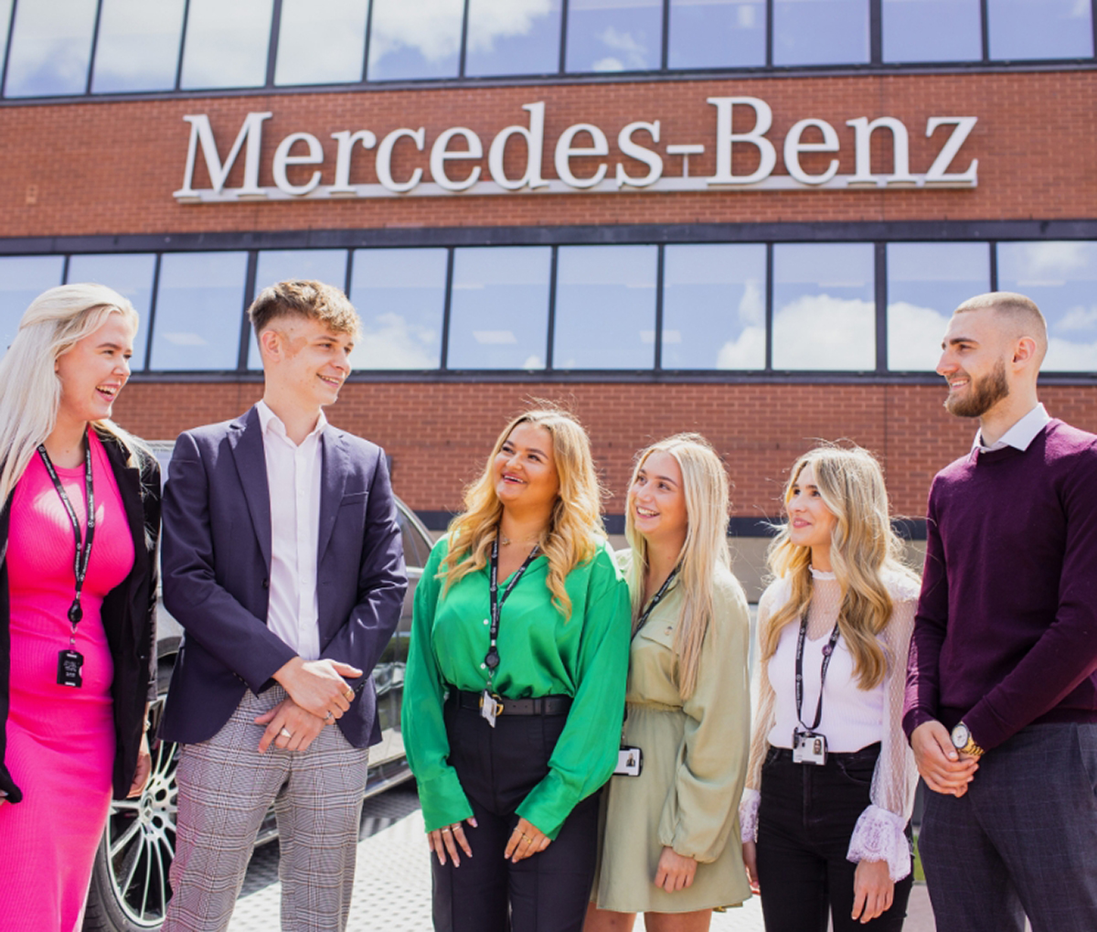 A group of people talking outside a building with a 'Mercedez-Benz' sign on the wall